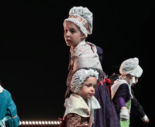 Des enfants vêtus de costumes d'époque, avec des bonnets blancs traditionnels, participent à une performance ou une scène historique.
