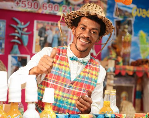 Vendeur souriant portant un chapeau en paille et un gilet coloré, montrant des boissons et produits sur son stand coloré, avec un fond de décor festif