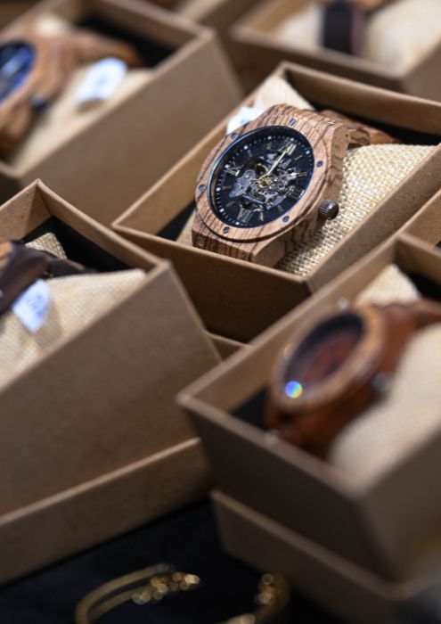 montres en bois de différentes formes et couleurs, présentées dans des boîtes en carton sur un stand.