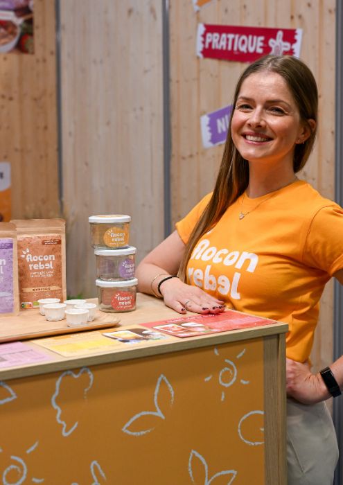 Exposante souriante devant son stand présentant des produits "Flocon Rebel", avec des emballages colorés et des échantillons à tester.