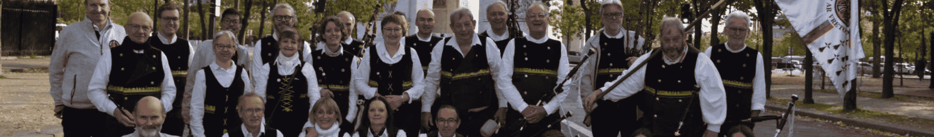 Un groupe d’hommes et de femmes, bretons, posent en plein air dans un parc boisé, vêtus de costumes traditionnels noirs et blancs, certains tenant des cornemuses.
