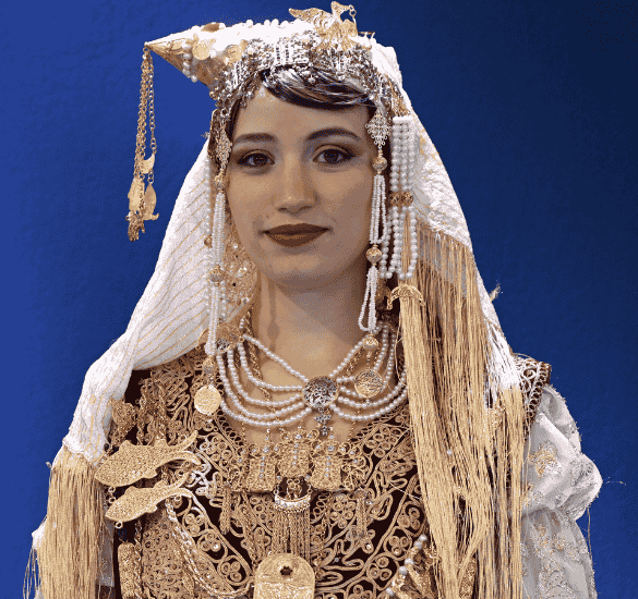 Femme portant un costume traditionnel richement brodé avec coiffe ornée de perles et bijoux dorés sur fond bleu.