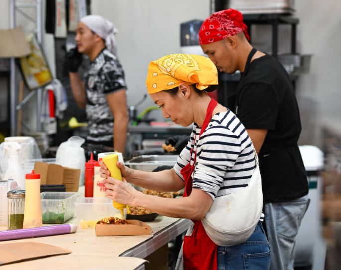 femme asiatique portant une mariniere, un bandana jaune et un tablier rouge en train de préparer un plat à emporter et d'appliquer de la moutarde dessus
