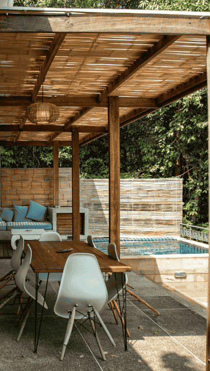Terrasse extérieure en bois avec pergola, grande table à manger, chaises blanches et piscine attenante entourée de végétation.