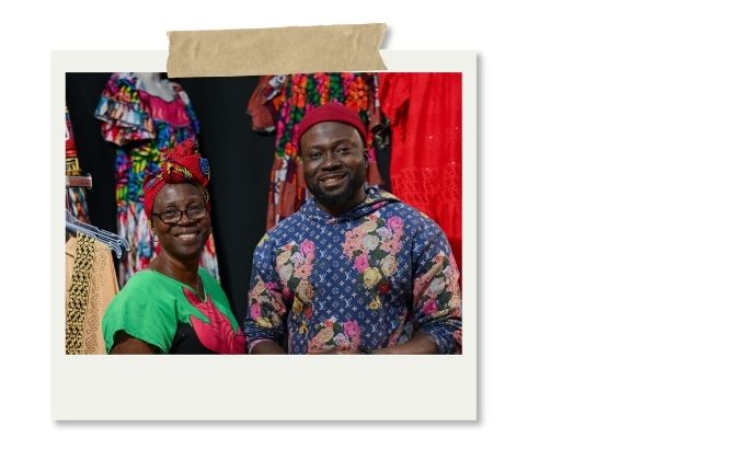 Deux personnes souriantes posent dans une boutique de vêtements colorés, entourées de tissus et tenues aux motifs africains.