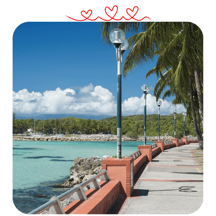 Une promenade pavée longe une mer turquoise, bordée de palmiers et de lampadaires, sous un ciel bleu avec des nuages.