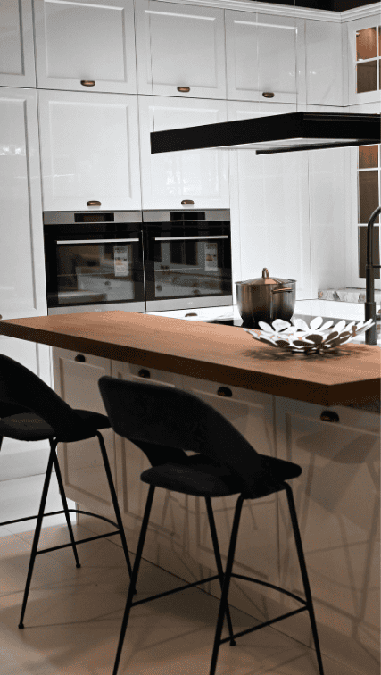 Cuisine moderne blanche avec ilot central en bois, tabourets noirs et fours encastres.