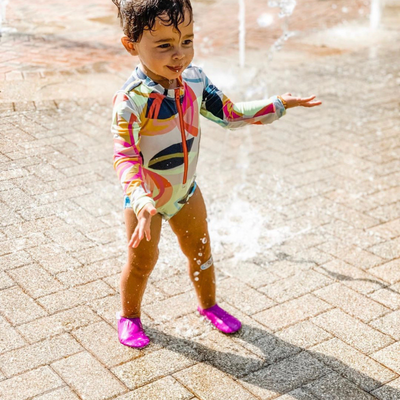 Petite fille en maillot de bain coloré jouant dans une fontaine d’eau sur un sol pavé en été.
