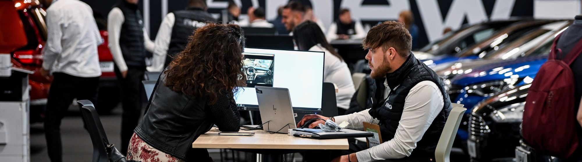 Deux personnes assises à une table de travail pendant une exposition automobile, analysant des photos de véhicule sur un écran d'ordinateur, entourées de voitures exposées