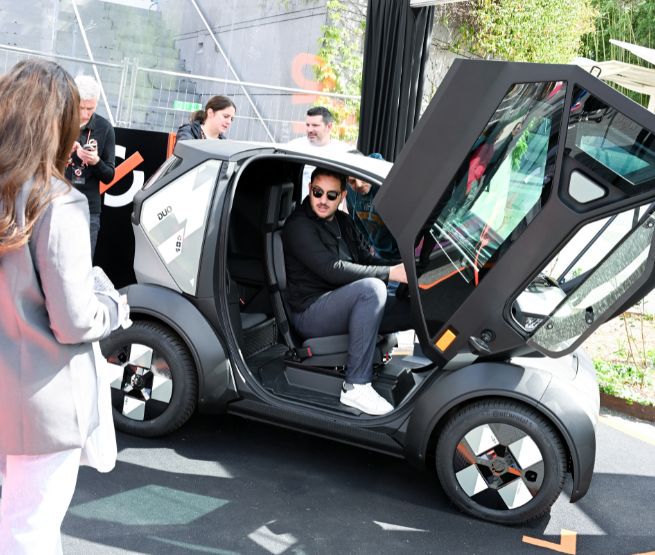 Visiteurs testant un véhicule électrique "Duo" lors d'une exposition automobile, avec un modèle à portes déployées