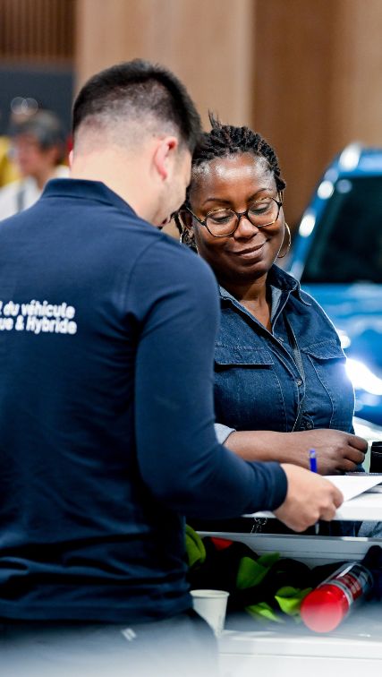 Deux personnes échangeant des informations sur les aides pour l'acquisition de véhicules électriques, avec des brochures et documents en fond