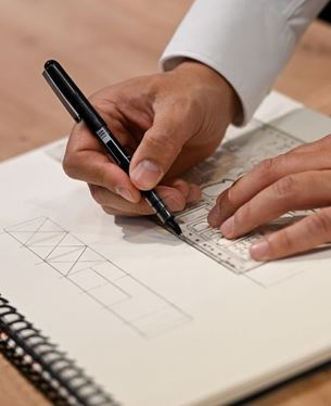 Gros plan sur des mains traçant un croquis technique au stylo et à la règle sur un carnet de dessin.