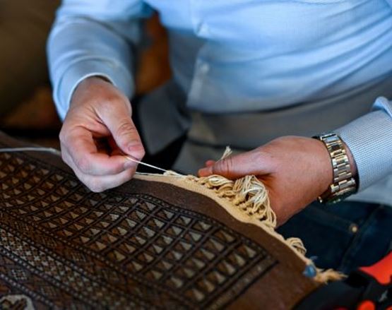 Mains d’un artisan cousant à la main un textile épais sur un établi, illustrant un savoir-faire traditionnel.